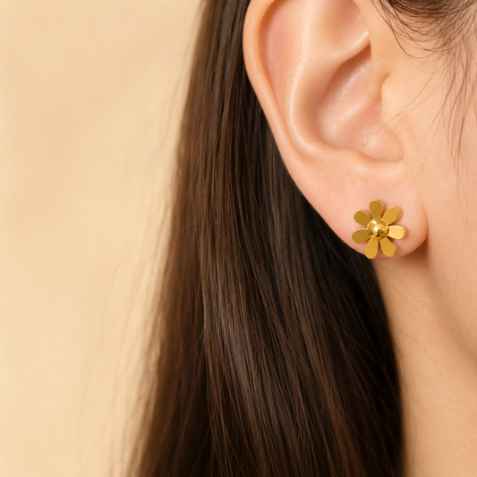 A close-up of a person's ear wearing a small gold flower stud earring against a soft beige background.