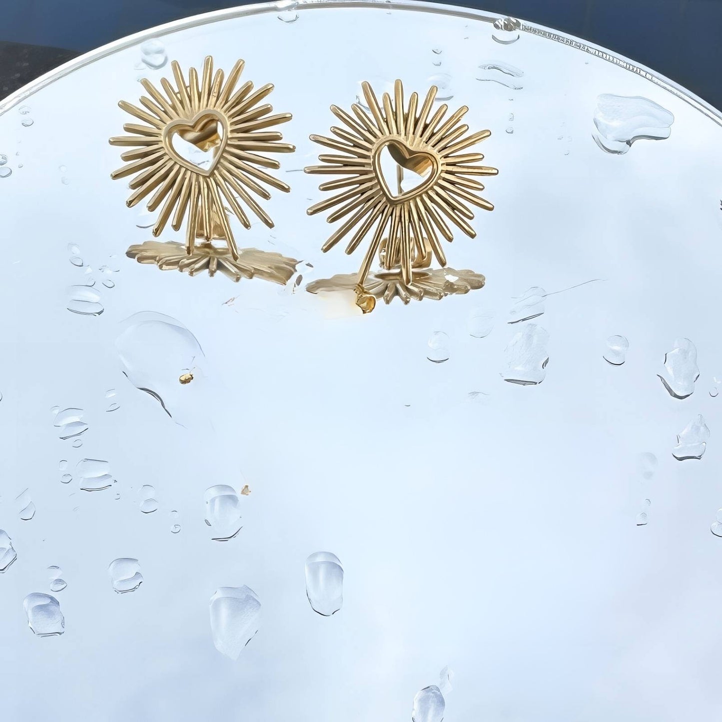 Gold sunburst earrings on a clear plastic stand with water droplets.
