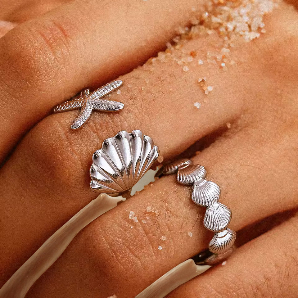 Silver starfish and shell rings on a person's fingers with sand in the background