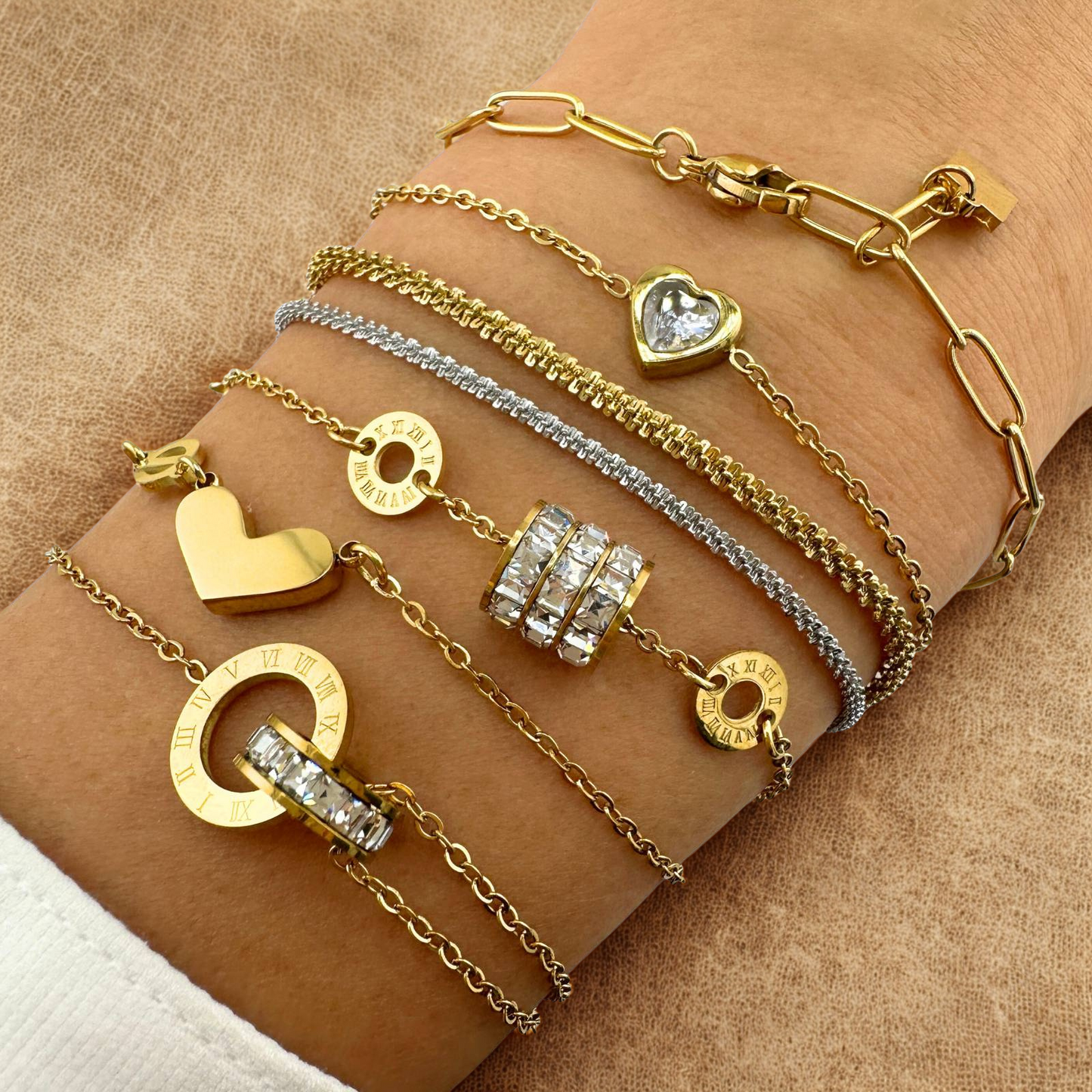 Gold and silver bracelets with heart and circular charms on a wrist against a beige background