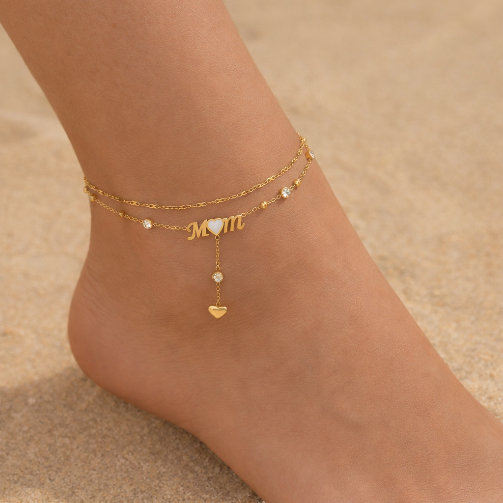 Gold anklets with 'MOM' and heart charms on a sandy background
