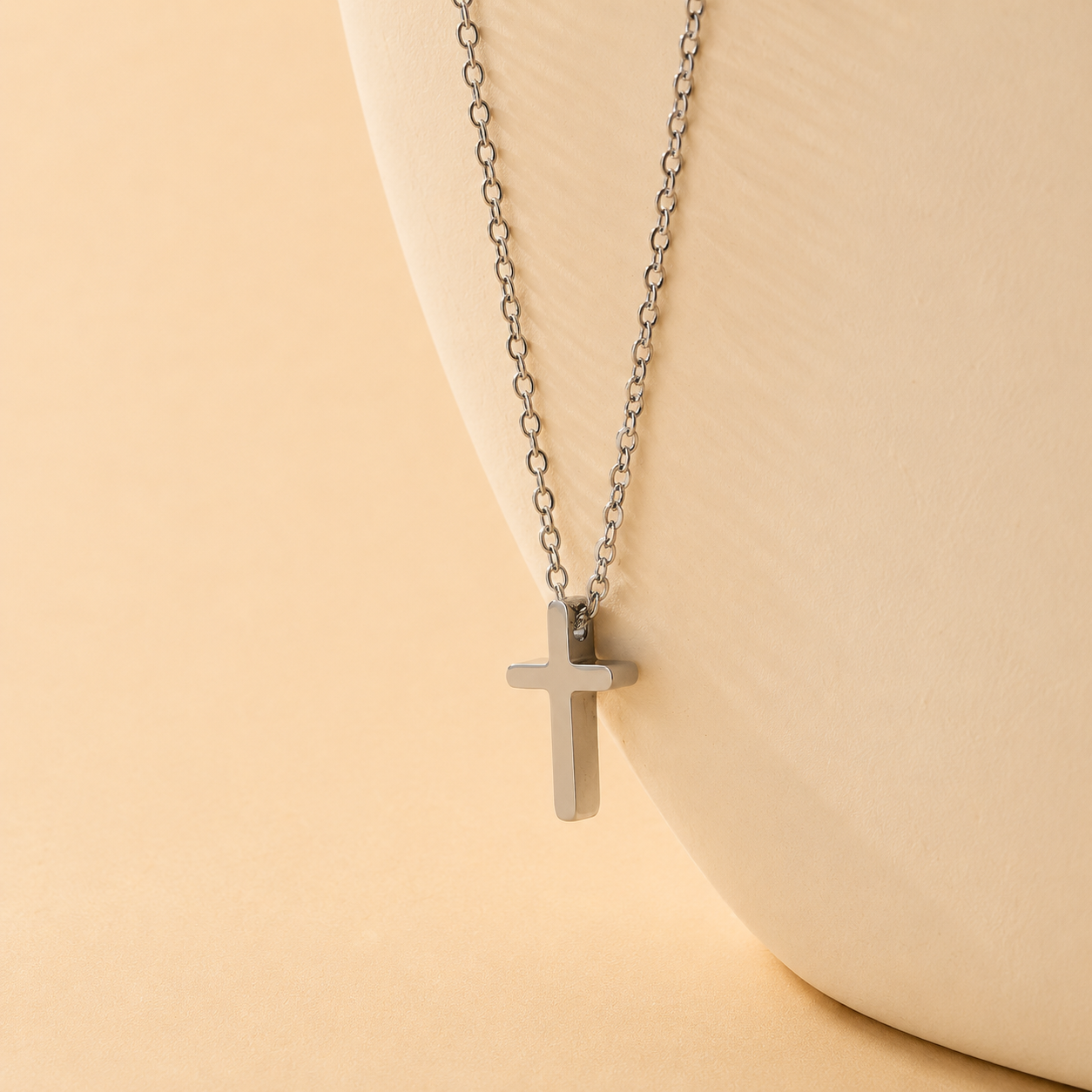 A silver cross pendant necklace on a delicate silver link chain, displayed on a curved cream-colored ceramic bust against a warm beige background.