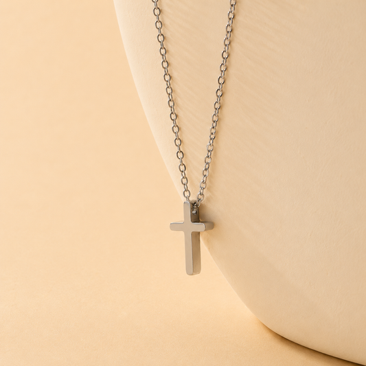 A silver cross pendant necklace on a delicate silver link chain, displayed on a curved cream-colored ceramic bust against a warm beige background.