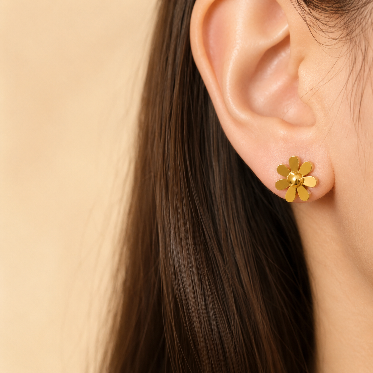 A close-up of a person's ear wearing a small gold flower stud earring against a soft beige background.