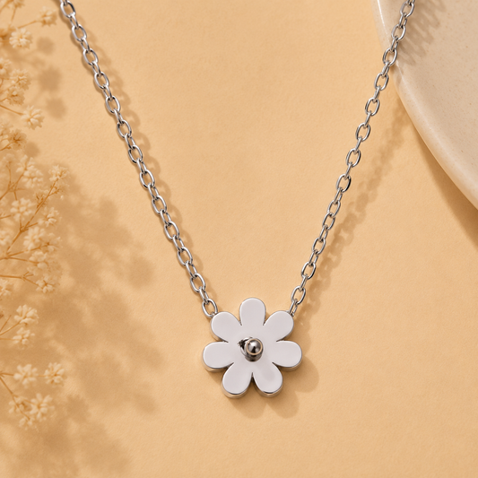 A silver flower pendant necklace with a five-petal design and a small central bead, displayed on a warm beige background with dried floral accents.
