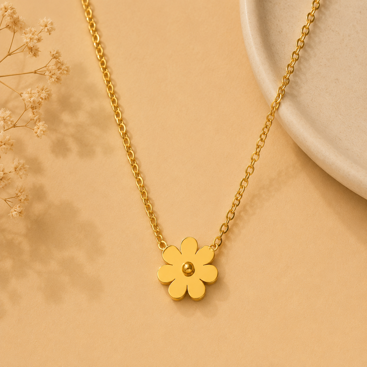 A gold flower pendant necklace with a small central bead, displayed on a warm beige background with dried floral accents and a ceramic plate edge.