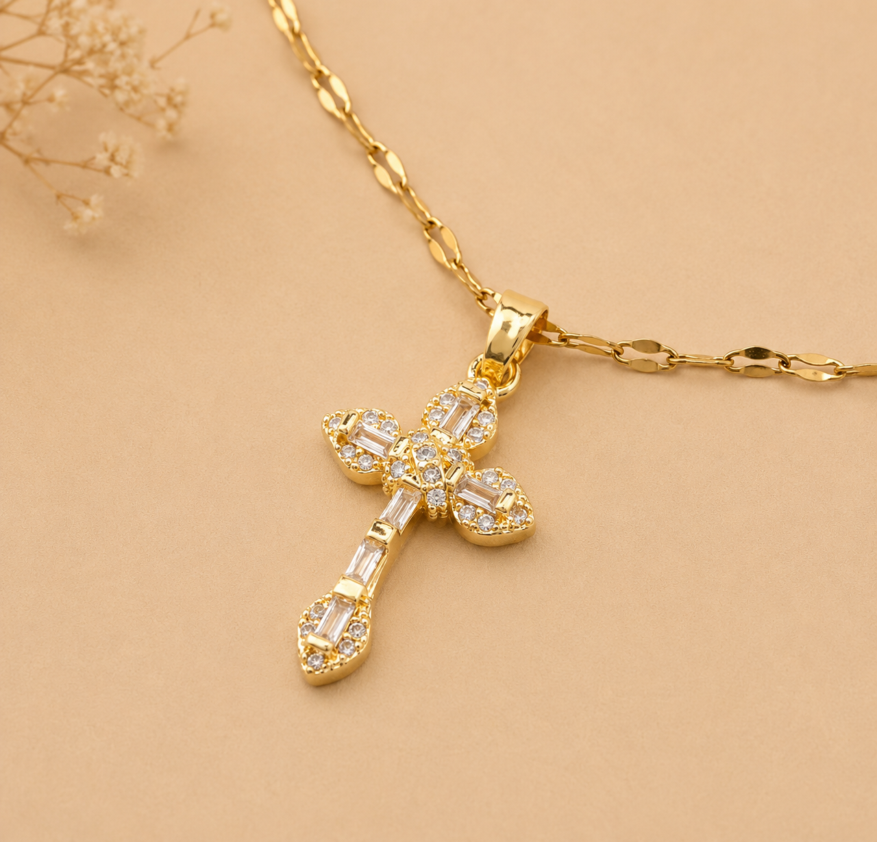 A gold cross pendant necklace adorned with clear baguette and round crystals, resting on a gold chain against a warm beige background with dried flowers in the corner.