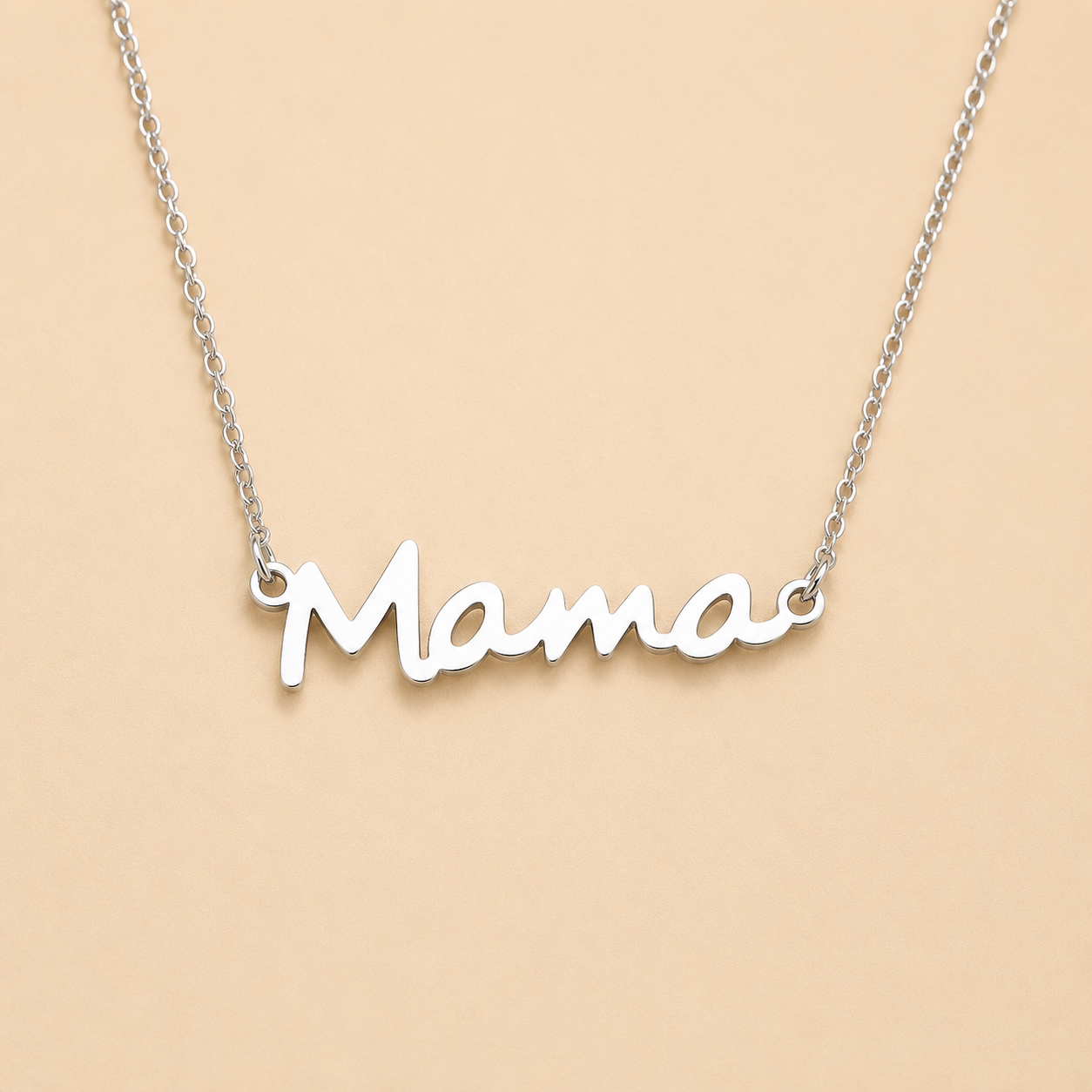 A silver nameplate necklace spelling 'Mama' in cursive script, suspended from a delicate silver chain against a solid light peach-beige background.