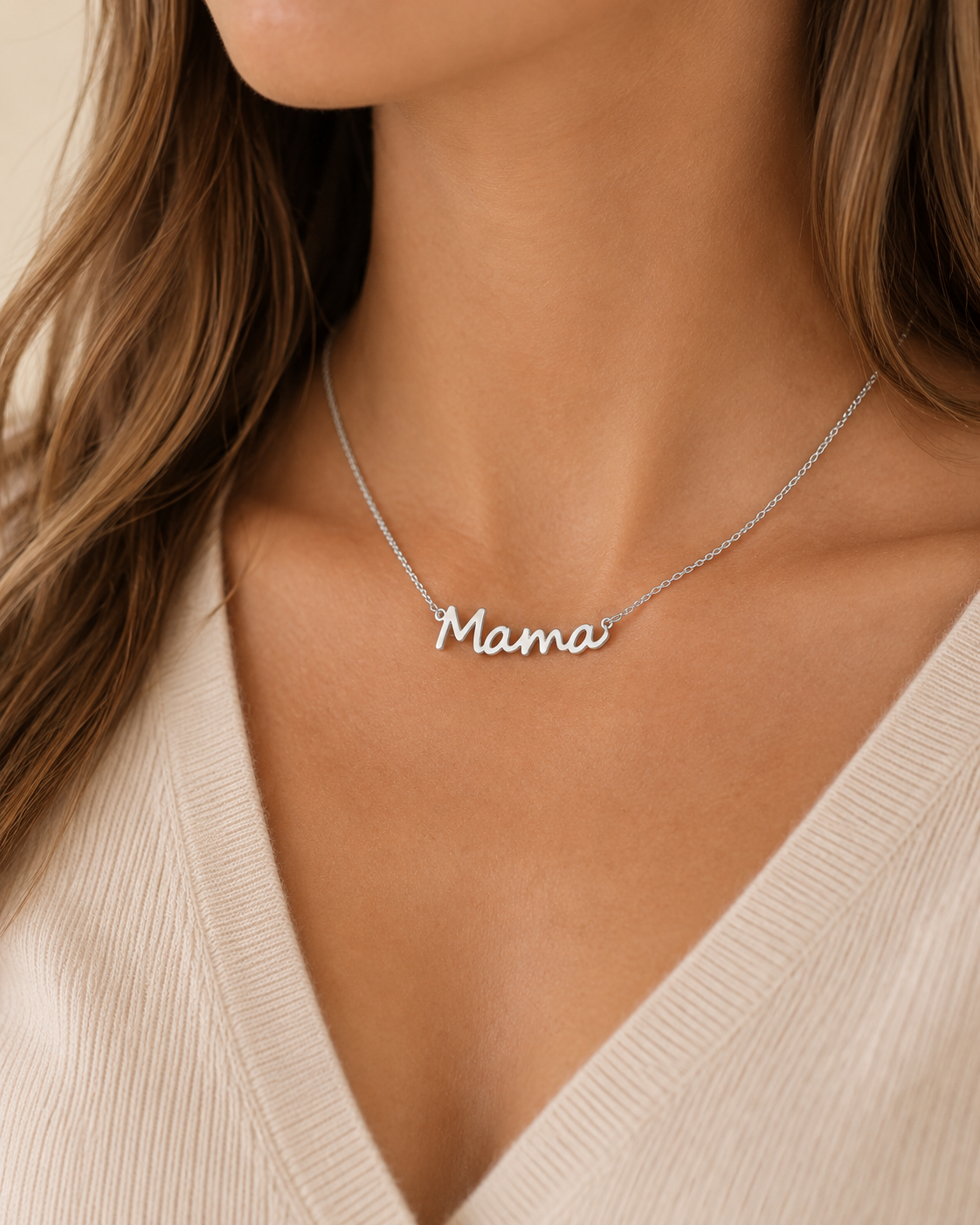 A close-up of a woman wearing a delicate silver script necklace that reads 'Mama' on a cream ribbed V-neck top.