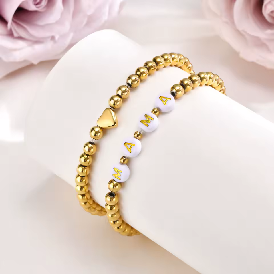 Gold beaded bracelet with 'MAMA' on a white surface with pink flowers in the background