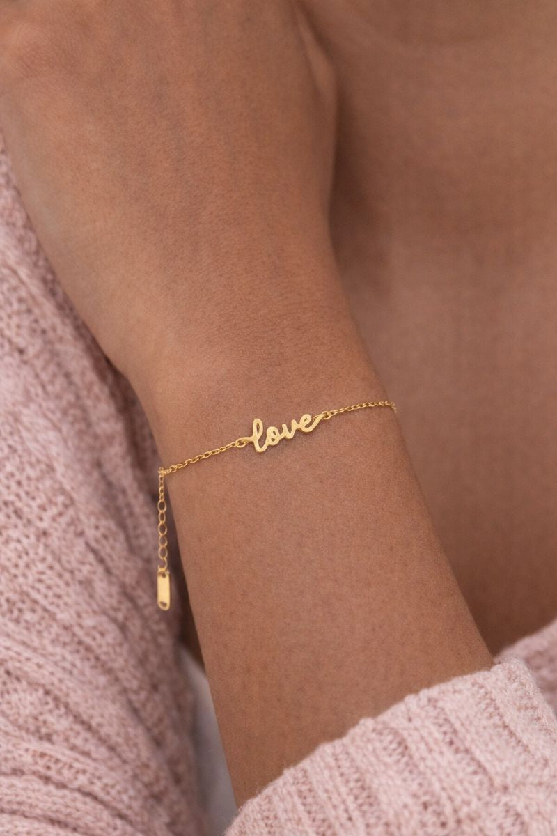 Gold 'love' bracelet on a wrist with a soft pink background