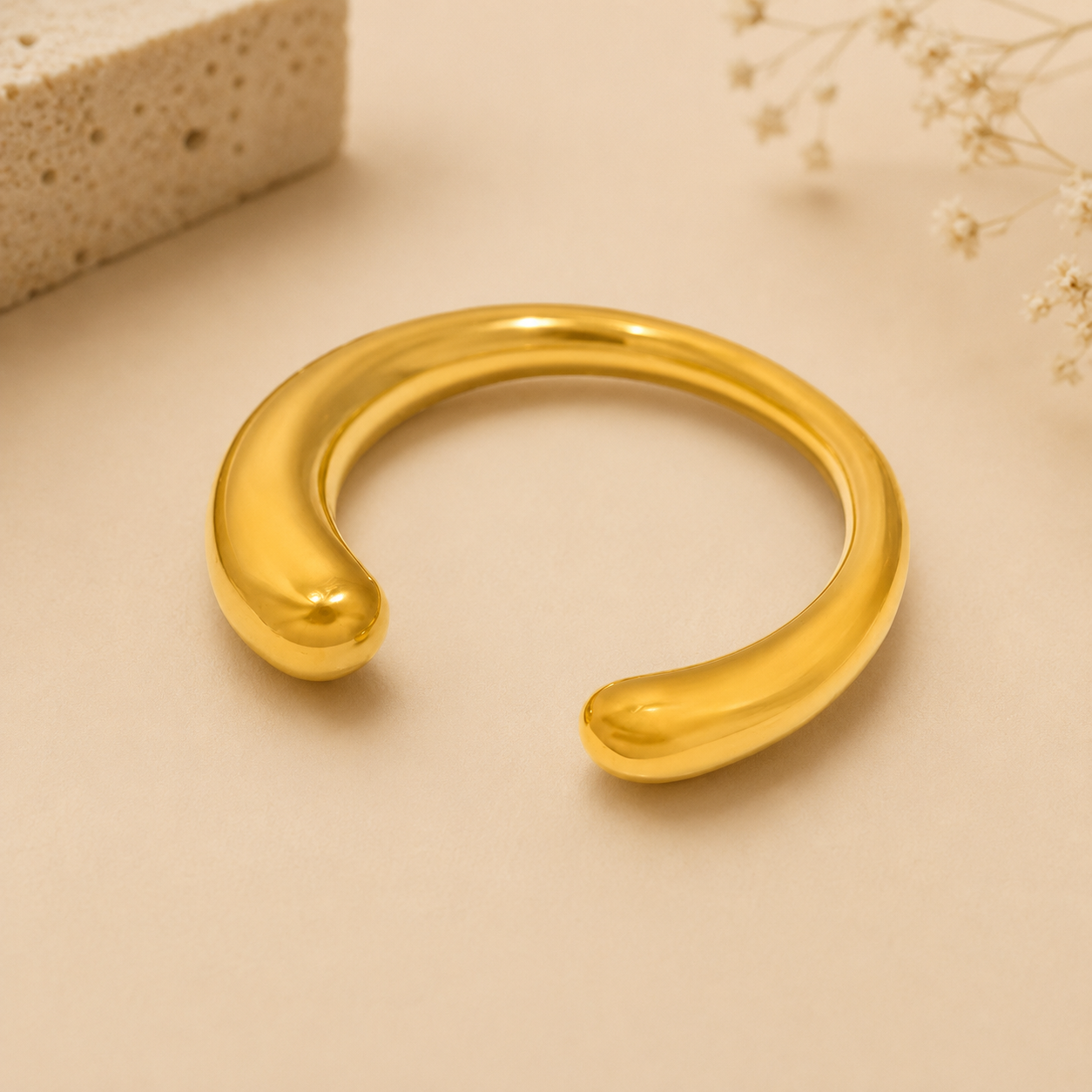 A polished gold-toned open cuff bracelet with rounded ends, displayed on a warm beige surface with soft decorative props in the corners.