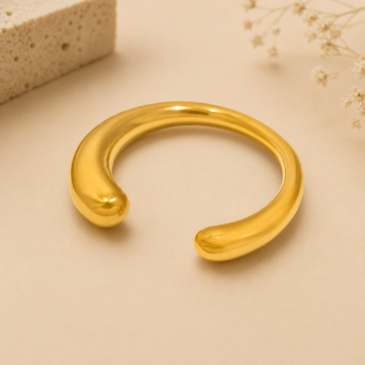 A polished gold-toned open cuff bracelet with rounded ends, displayed on a warm beige surface with soft decorative props in the corners.