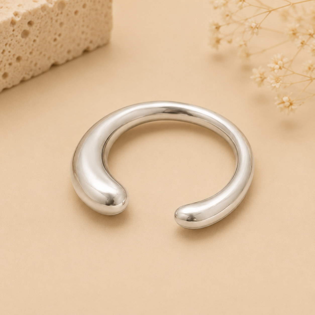 A polished silver-toned open cuff bracelet with rounded ends, displayed on a warm beige surface with dried flowers and a textured stone prop in the background.