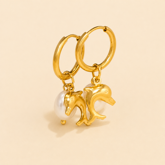 A pair of gold hoop earrings with dangling dolphin charms and a white pearl, shown on a solid light beige background.