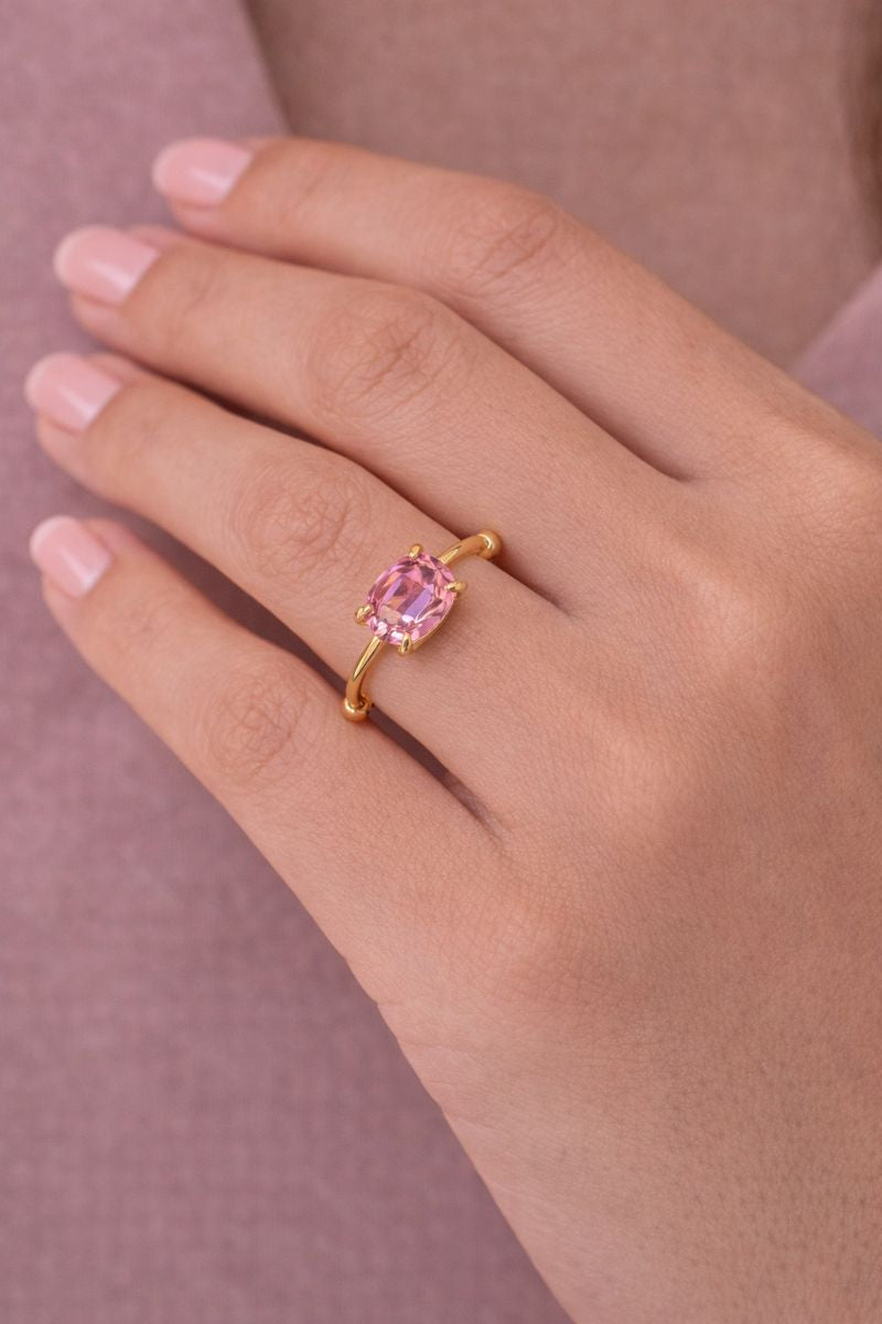 Gold ring with a pink gemstone on a hand against a pink background