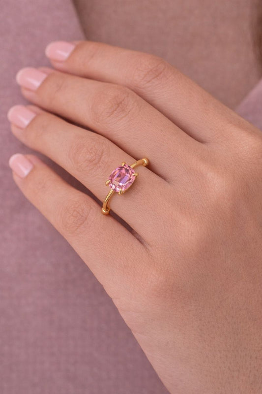 Gold ring with a pink gemstone on a hand against a pink background